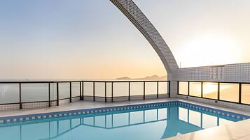 A swimming pool with a curved glass barrier, a clear view of the sea, and a sunset in the background.