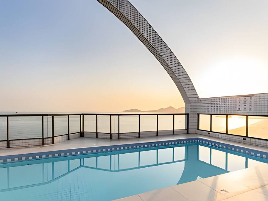 Een zwembad met een gebogen glazen barrière, uitzicht op zee en een zonsondergang op de achtergrond.