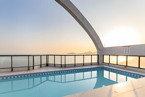 Ein Schwimmbad mit einer gewölbten Glasscheibe, freiem Blick auf das Meer und einem Sonnenuntergang im Hintergrund.
