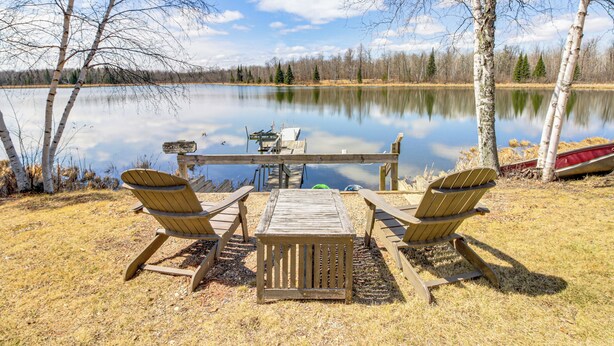 Eine Holzterrasse mit zwei Adirondack-Stühlen und einem Picknicktisch mit Blick auf einen ruhigen See, umgeben von Birken.
