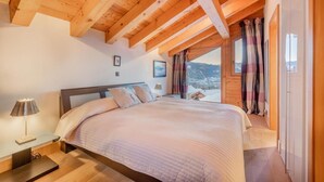 Un dormitorio con techo de madera, una cama con colcha blanca, una mesita de noche con lámpara y una ventana con cortinas.