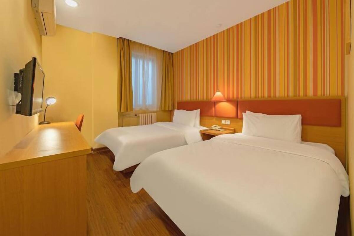 A hotel room with two beds, a wooden desk, a mirror, a lamp, and a window with curtains.
