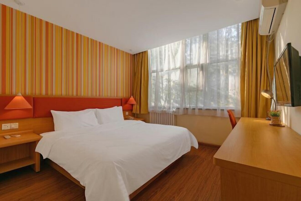 A hotel room with a bed, a desk, a chair, a nightstand, a lamp, a TV, and a window with curtains.