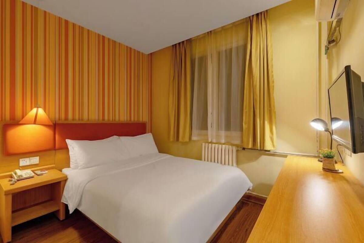 A hotel room with a bed, bedside table, lamp, window with curtains, and a TV on the wall.