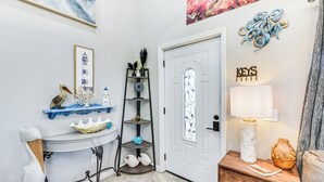 Um corredor bem iluminado com uma porta branca, uma prateleira com objetos decorativos, uma mesa com um vaso e uma parede com um quadro emoldurado e uma placa.