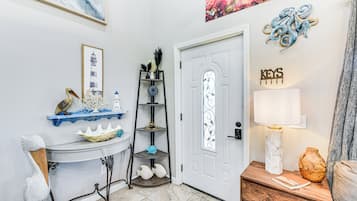 A well-lit hallway with a white door, a shelf with decorative items, a table with a vase, and a wall with a framed picture and a sign.