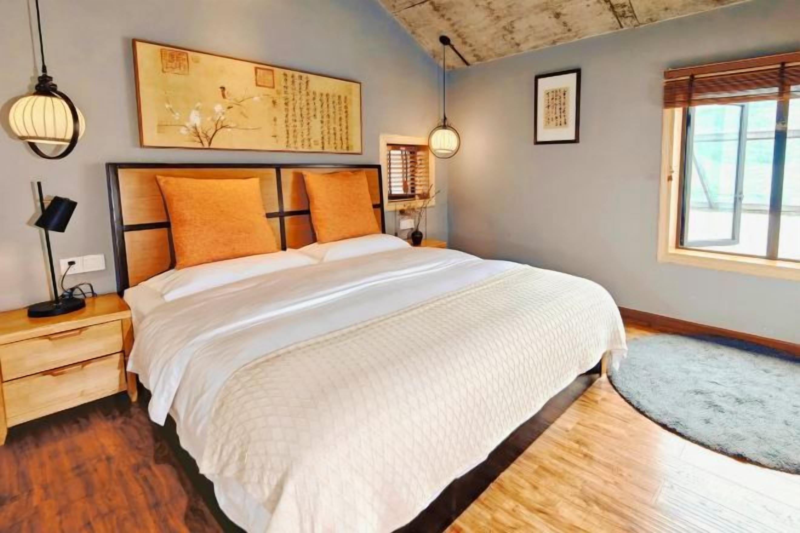 A bedroom with a large bed, wooden headboard, two bedside tables, a nightstand, a window with blinds, and a wall-mounted artwork.