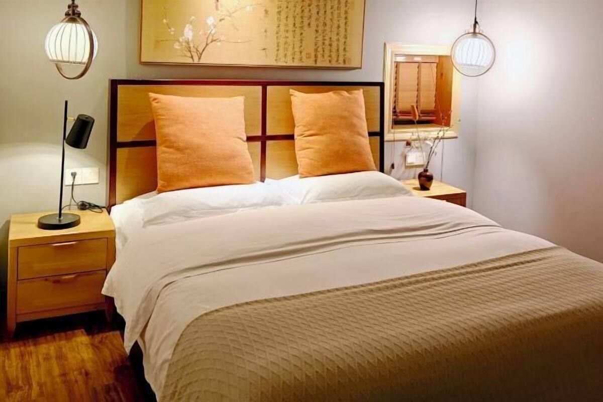 A neatly made bed with a headboard, two bedside tables with lamps, a wall-mounted artwork, and a window with blinds.