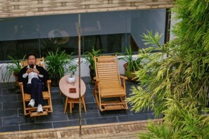 Une personne est assise sur une chaise en bois, à l’extérieur d’un bâtiment doté d’une porte en verre, avec une petite table ronde et des plantes en pot.