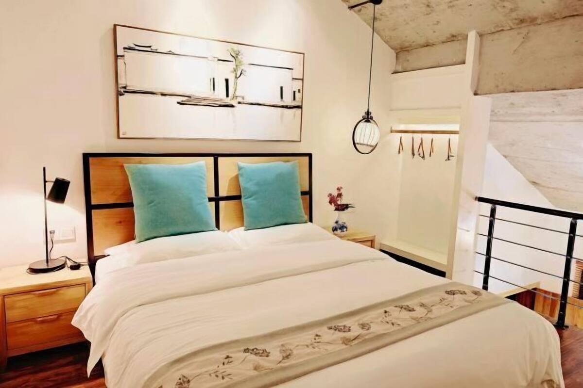 A bedroom with a bed, two nightstands, a wall-mounted artwork, a hanging light fixture, and a window with a view of a balcony.