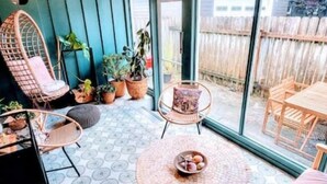 Eine gemütliche Terrasse mit rundem Tisch, zwei Stühlen und einer Hängematte. Im Hintergrund befinden sich mehrere Kübelpflanzen und ein Holzzaun.