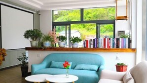 A room with a teal sofa, a white table, and a bookshelf filled with books. There are potted plants on a shelf above the sofa and a window with a view of greenery. - Moment Inn (Huangshan City)