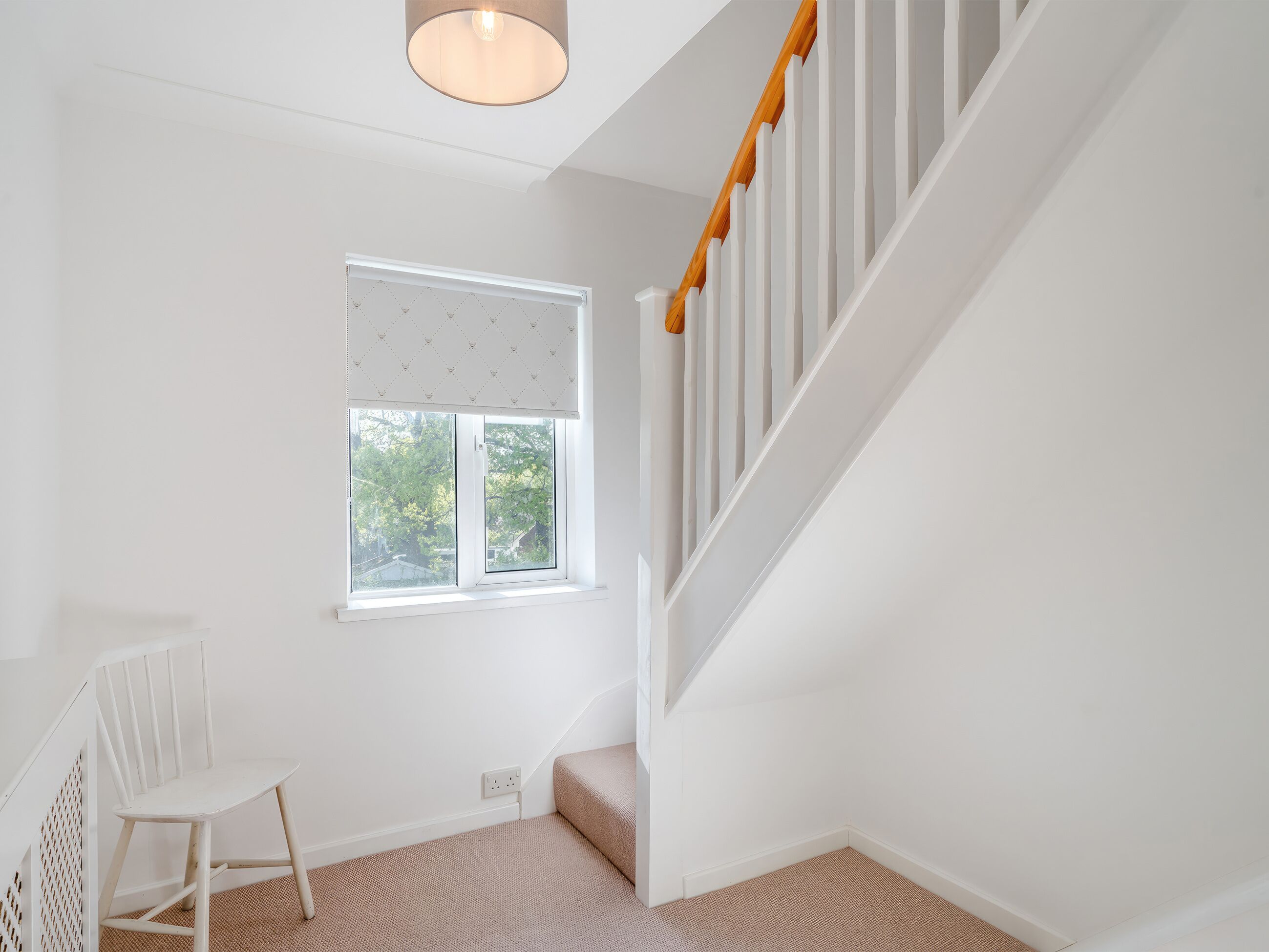 A room with a white wall, a window with blinds, a white chair, a staircase with wooden railings, and a carpeted floor.