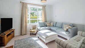 A living room with a sectional sofa, a small armchair, a television on a wooden stand, and a large window with curtains. - 5 bedroom accommodation in Bransgore, near Christchurch (Bransgore, near Christchurch)