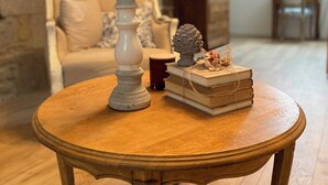 Una mesa redonda de madera con un candelabro y libros, frente a una pared de piedra y una silla blanca en una habitación con suelo de madera.