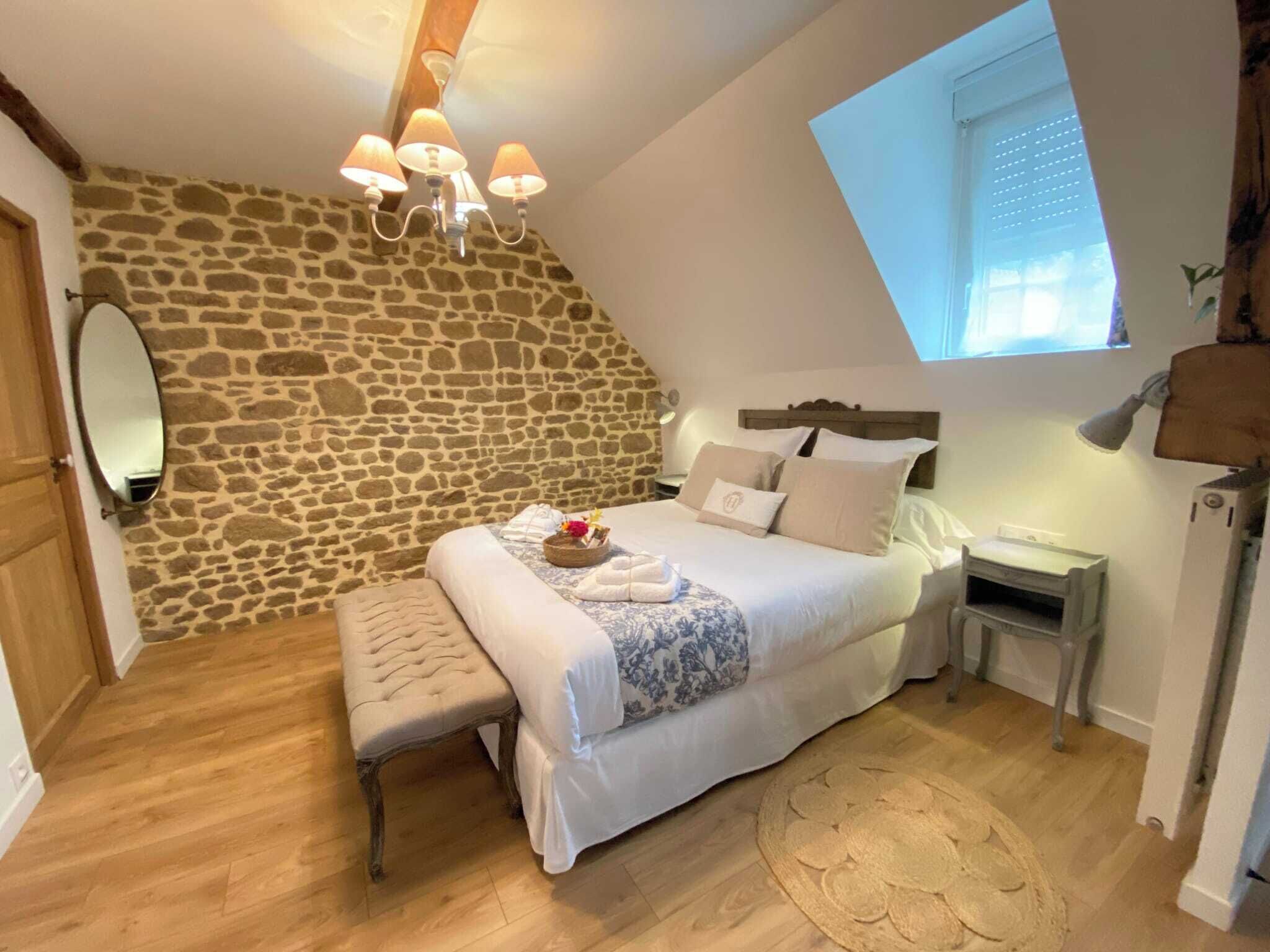 A bedroom with a stone wall, a bed with white linens, a bench, a bedside table, and a window with blinds.