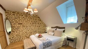 Un dormitorio con pared de piedra, una cama con ropa blanca, una banca, una mesita de noche y una ventana con persianas.