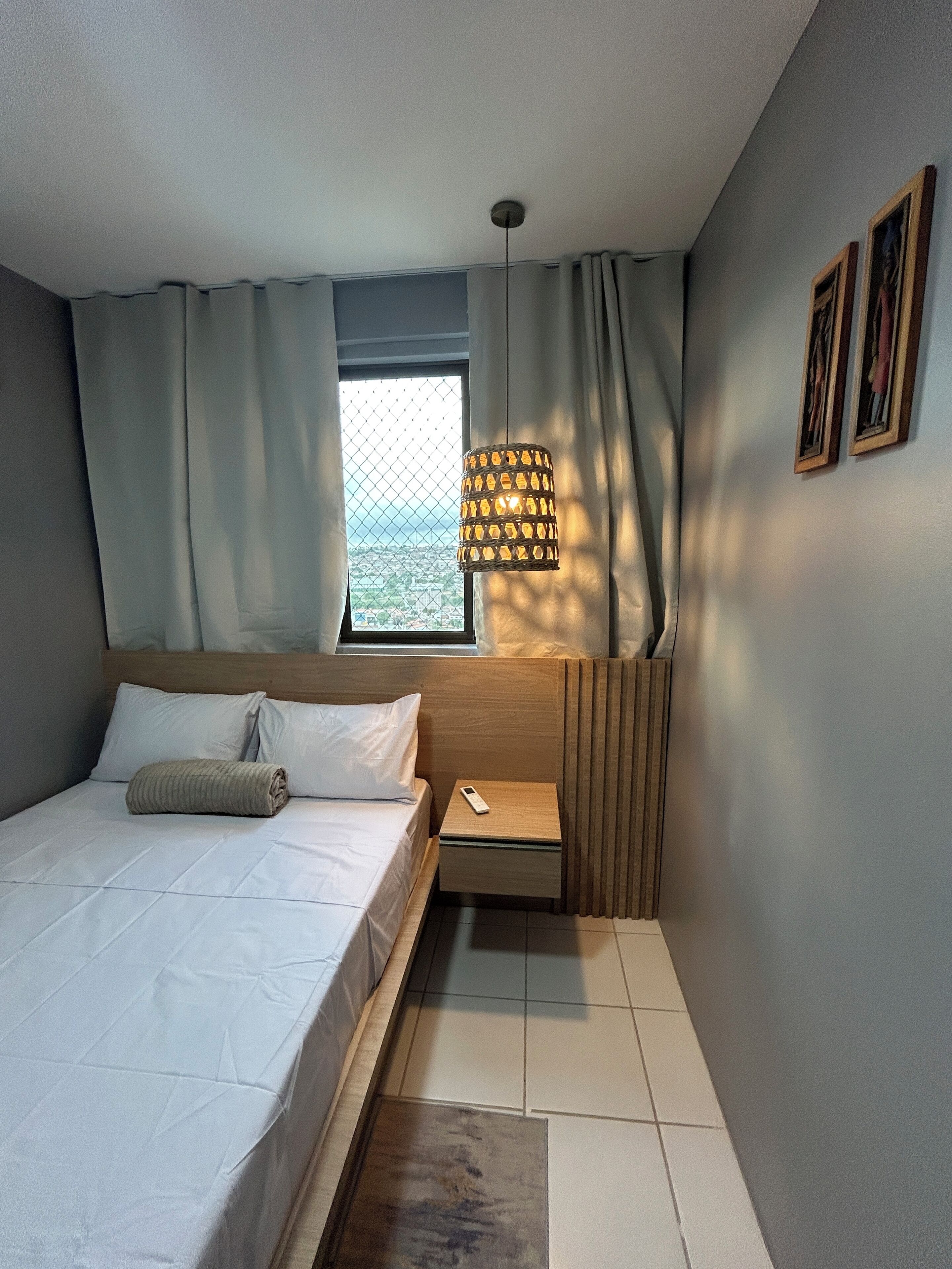 A bedroom with a bed, a bedside table, a hanging lamp, a window with curtains, and a view of the outside.