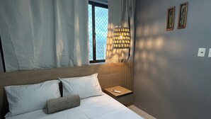 A neatly made bed with white linens, a wooden headboard, and two rolled towels. There is a bedside table with a lamp and a remote control. The room has a window with curtains and a view of the outside.
