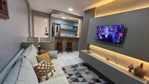 Een moderne woonkamer met een bank, een aan de muur bevestigde televisie en een keuken op de achtergrond.