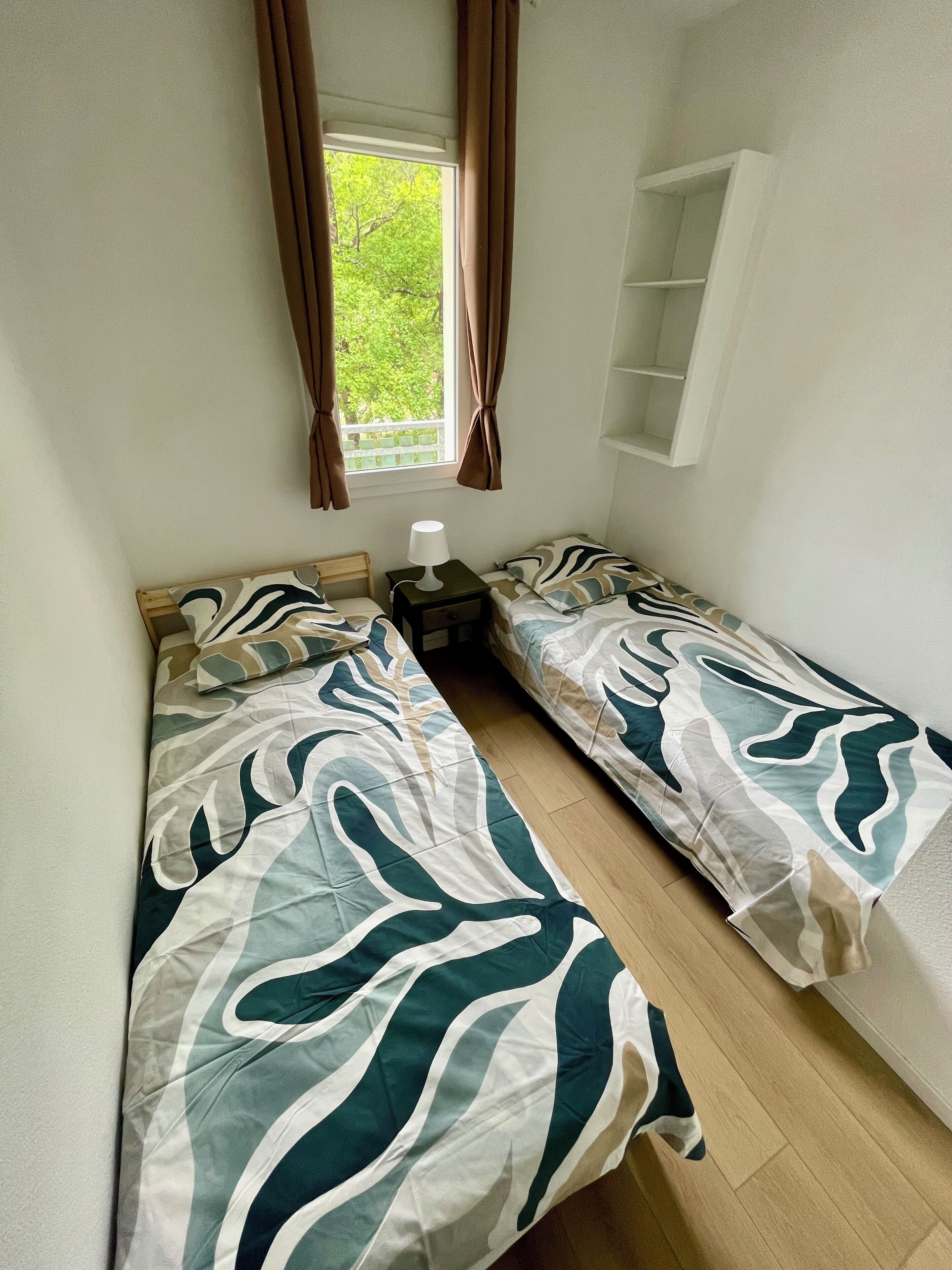 Une chambre avec deux lits simples, une table de chevet avec une lampe, une fenêtre avec des rideaux et une étagère blanche.