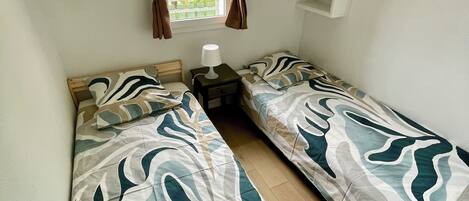Une chambre avec deux lits simples, une table de chevet avec une lampe, une fenêtre avec des rideaux et une étagère blanche.