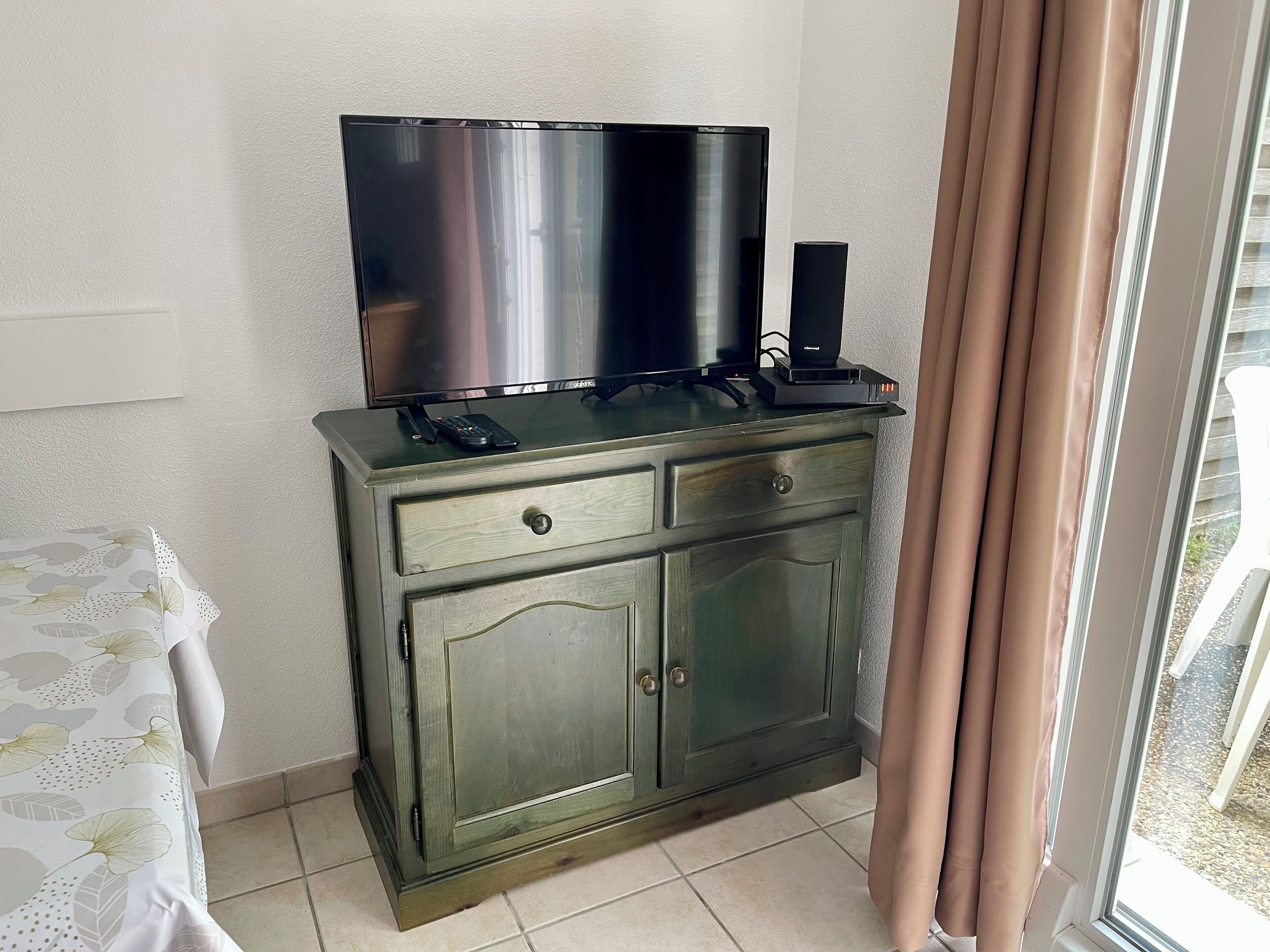 Une pièce avec un téléviseur posé sur un meuble, une petite table recouverte d’une nappe à motifs floraux et une fenêtre avec des rideaux.