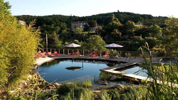 Zona de piscina con sillones de descanso y sombrillas, rodeada de vegetación y colinas al fondo.