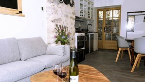 Una sala de estar moderna con un sofá gris, una mesa de centro de madera con una botella de vino y copas, una planta decorativa y una cocina al fondo.