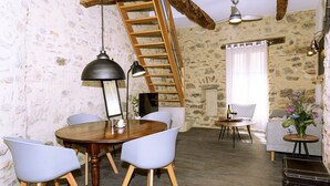 Una habitación rústica con una mesa de comedor de madera, sillas blancas, una escalera, un sofá y una pequeña mesita con un jarrón.