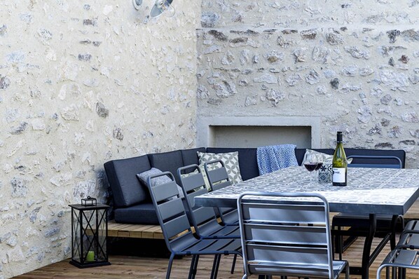 Un espace extérieur aménagé pour les repas, avec un mur en pierre, un banc en bois, des chaises en métal et une table où est placée une bouteille de vin.