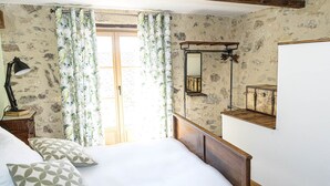 A bedroom with a wooden bed, a nightstand with a lamp, floral curtains, and a stone-patterned wall. - La Grange Droite Cottage 3 Stars (BELESTA)