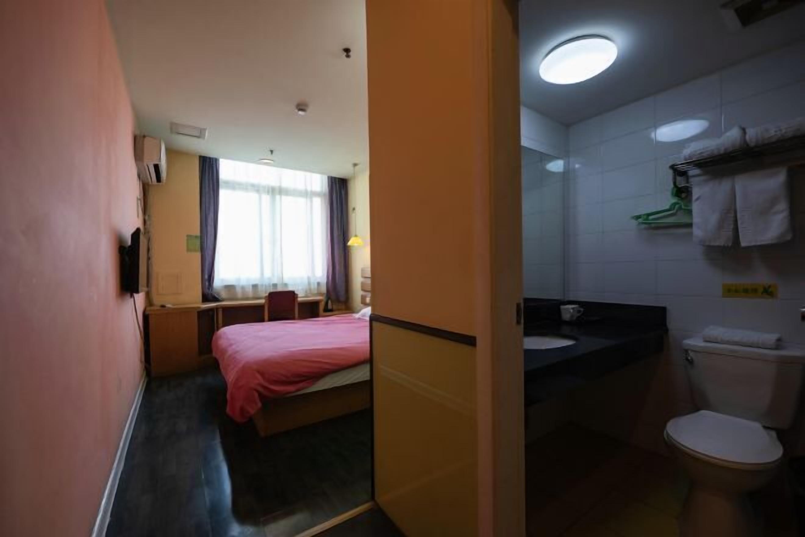 Ein Hotelzimmer mit Bett, kleinem Schreibtisch, Stuhl, Fenster mit Vorhängen sowie einem Badezimmer mit Toilette, Waschbecken und Handtuchhalter.