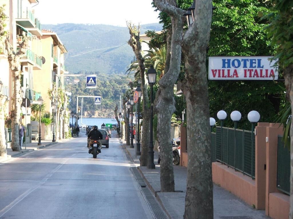Una strada con un cartello per HOTEL VILLA ITALIA, alberi lungo il marciapiede e un paesaggio collinare sullo sfondo.