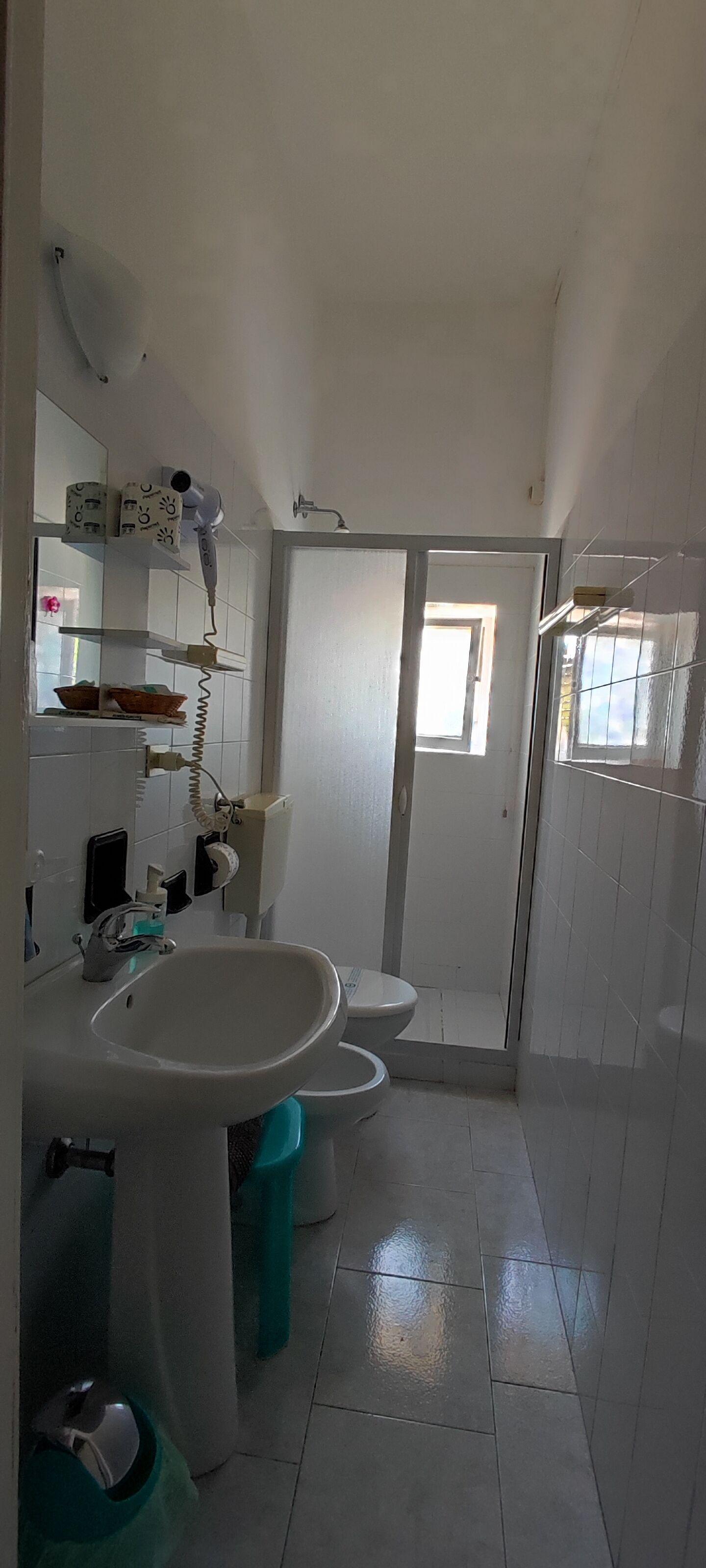 Une salle de bain avec un lavabo, des toilettes et une douche, le tout de couleur blanche. On y trouve des étagères avec des articles de toilette et un sèche-cheveux fixé au mur.