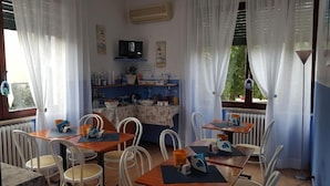Un espace repas avec des tables et des chaises, un téléviseur fixé au mur et un coin cuisine équipé d’étagères et d’appareils électroménagers.