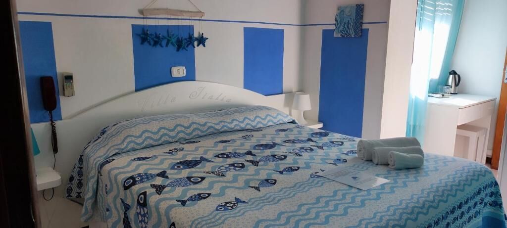 Un letto con copriletto a fantasia blu e bianca, un comodino con lampada e un telefono fissato alla parete.