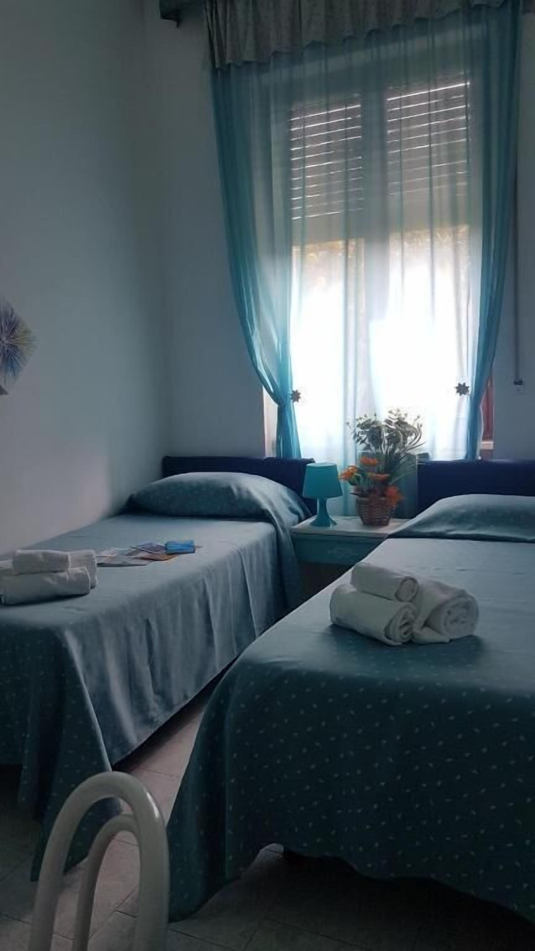 Una habitación con dos camas, una silla, un ventanal con cortinas y una mesita con lámpara y planta.