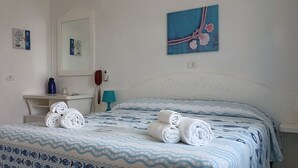 Un lit bien fait, avec des serviettes pliées, une table de chevet avec une lampe, un miroir et un mur orné d’un tableau encadré et d’une affiche.