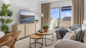 Un salon moderne doté d’un téléviseur à écran plat, d’un canapé, d’un coin repas avec des chaises en osier et offrant une vue sur le paysage urbain grâce à de grandes fenêtres.
