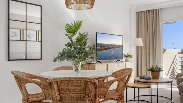 Un salon moderne avec une table à manger ronde, des chaises en osier, un grand téléviseur et une vue sur la mer.