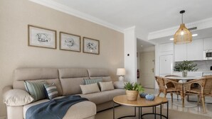 Un salon comprenant un canapé, une table basse ronde et un coin repas avec des chaises en osier. Des tableaux encadrés ornent le mur, et un luminaire suspendu éclaire la pièce.