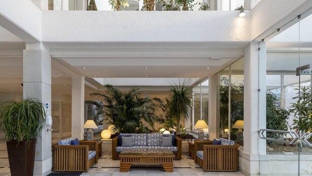 Un hall d’entrée spacieux avec un espace salon central, de grandes plantes en pot et un mur de verre donnant sur l’extérieur.