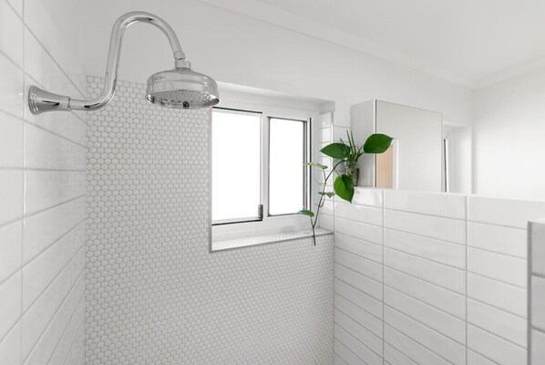 A bathroom with white tiled walls, a shower with a chrome handle, a window, and a plant.