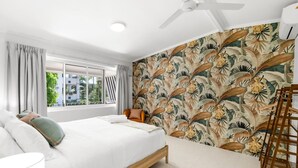 Un dormitorio con una cama, una silla, un ventanal y una pared con un estampado de hojas tropicales.