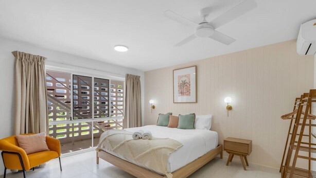 Un dormitorio con cama, una silla, una mesita de noche, un ventilador de techo y una ventana con vistas a un edificio.