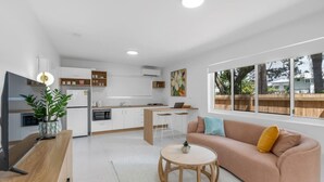 Una cocina moderna con armarios blancos, un comedor con mesa y sillas de madera, un sofá con cojines, y un amplio ventanal con vista a árboles.