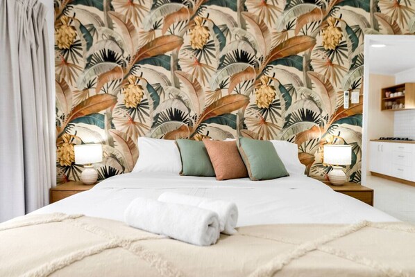 Una cama bien hecha con sábanas blancas, una toalla enrollada y varios almohadones de diferentes colores.