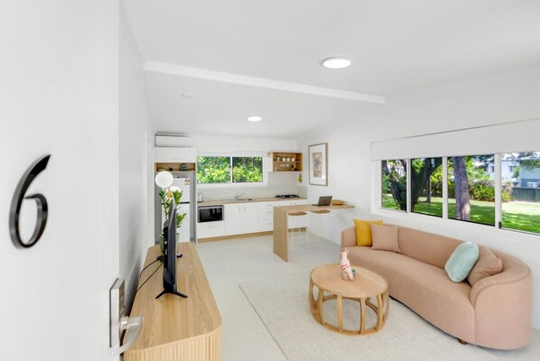 Un salon moderne et bien éclairé, avec un canapé, une table basse et un téléviseur. On aperçoit une cuisine à l’arrière-plan, avec une fenêtre donnant sur la verdure à l’extérieur.