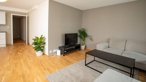 Un salon moderne avec un plancher en bois, un canapé gris, une table basse noire, un téléviseur posé sur un meuble et deux plantes en pot.